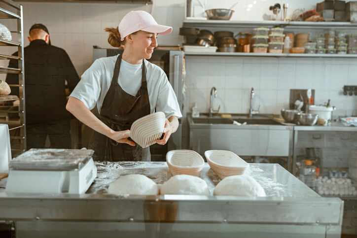 A bakery worker smiling and prepping loaf pans for bread in the kitchen.