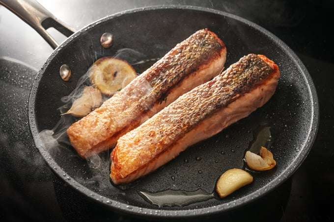 Two slices of fresh salmon fillet with a crispy fried skin in a black pan with garlic, thyme and lemon on the stove, cooking a delicious seafood meal, selected focus