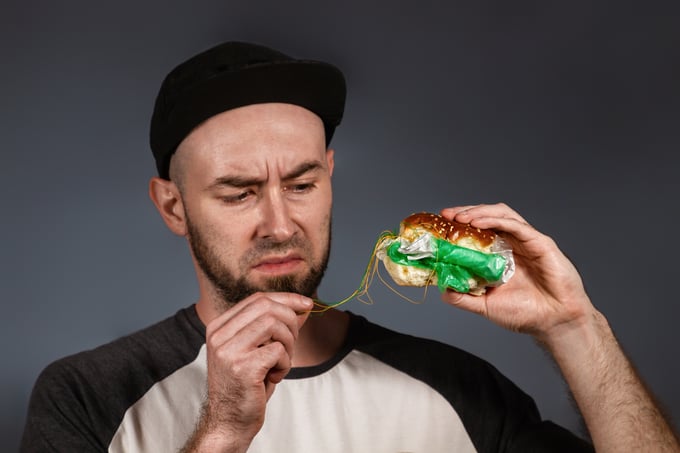 The concept of fast food and ecology. A man in a cap and a beard, holding a hamburger, and with disgust pulls out a thread. Black background