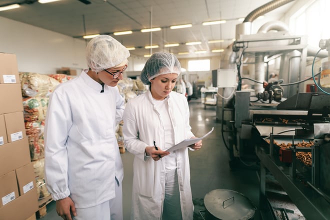 Two quality professionals in white sterile uniforms checking quality of salt sticks while standing in food factory
