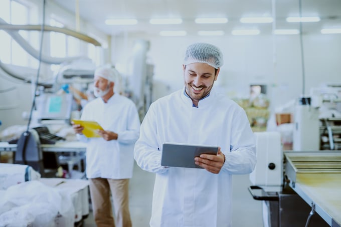 Young Caucasian smiling supervisor in sterile white uniform using tablet while standing in food plant